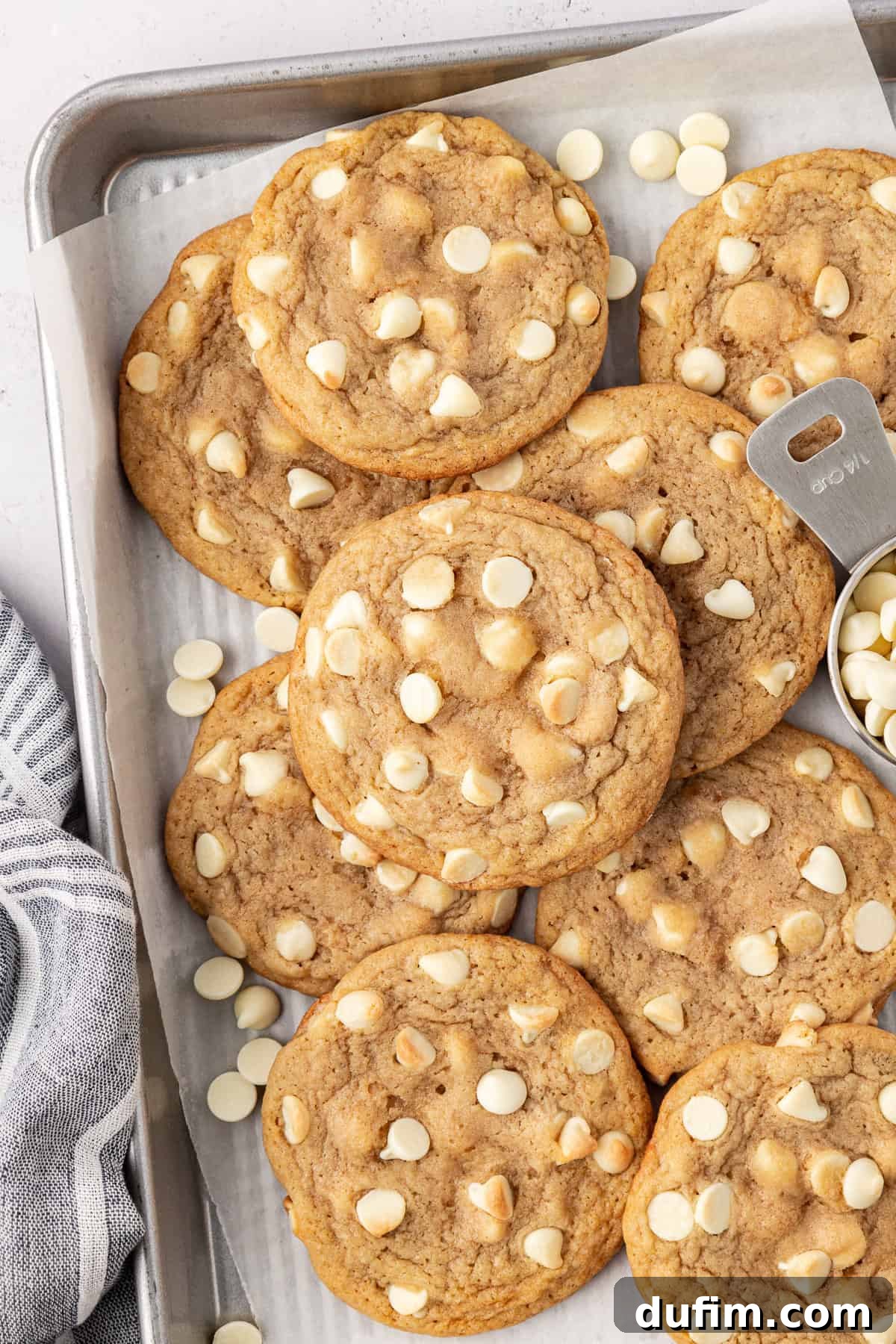 White chocolate chip cookies and white chocolate chips piled and scattered on a baking sheet lined with white parchment paper