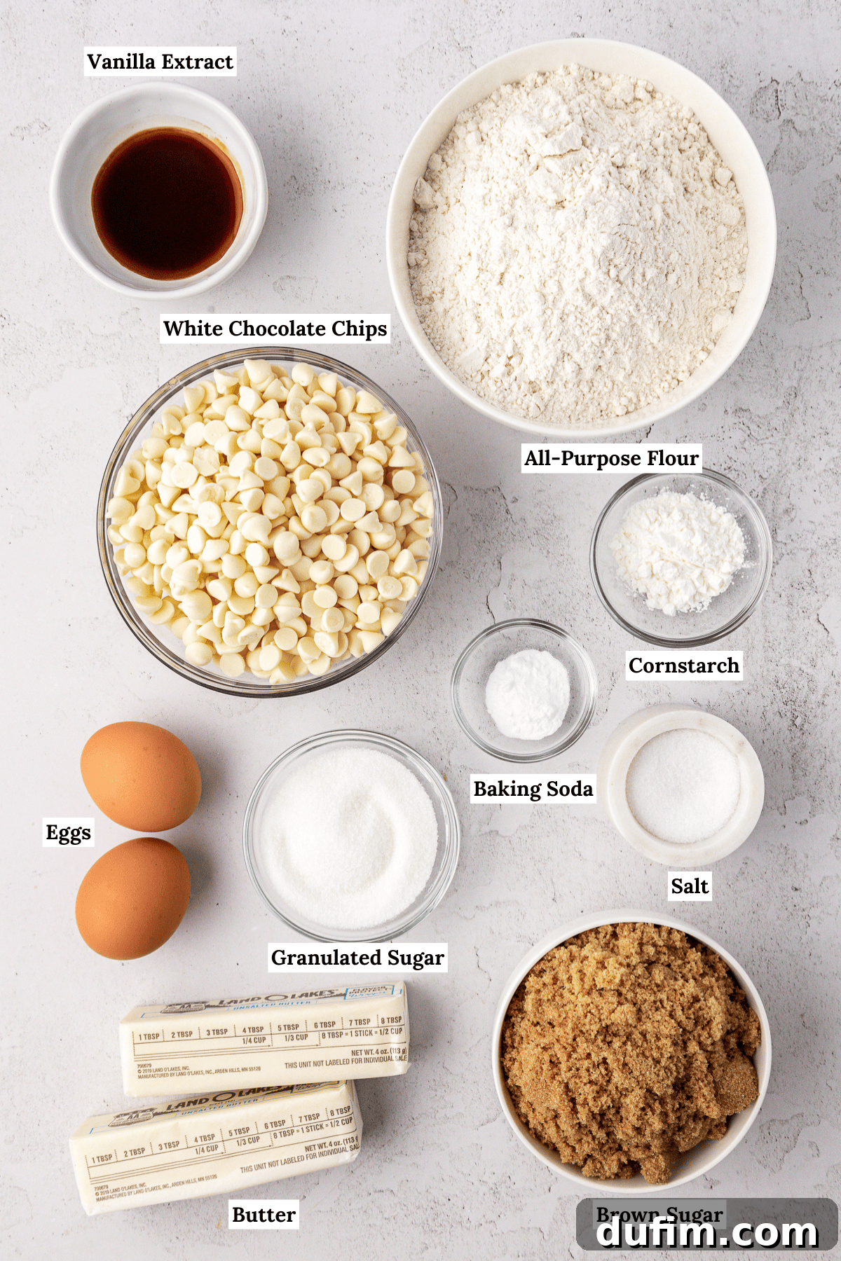Overhead view of white chocolate chip cookie ingredients including vanilla extract, all-purpose flour, white chocolate chips, cornstarch, eggs, baking soda, granulated sugar, salt, butter and brown sugar