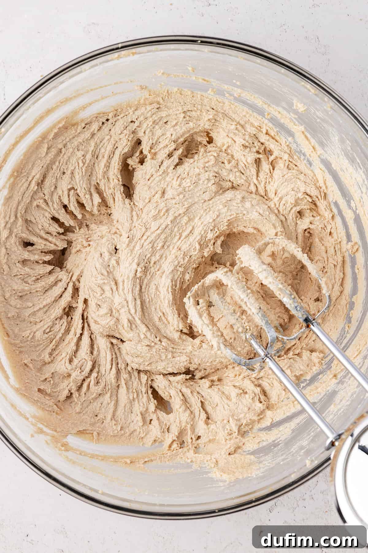 Cookie dough batter in a clear glass bowl with an electric mixer leaning on the side of the bowl