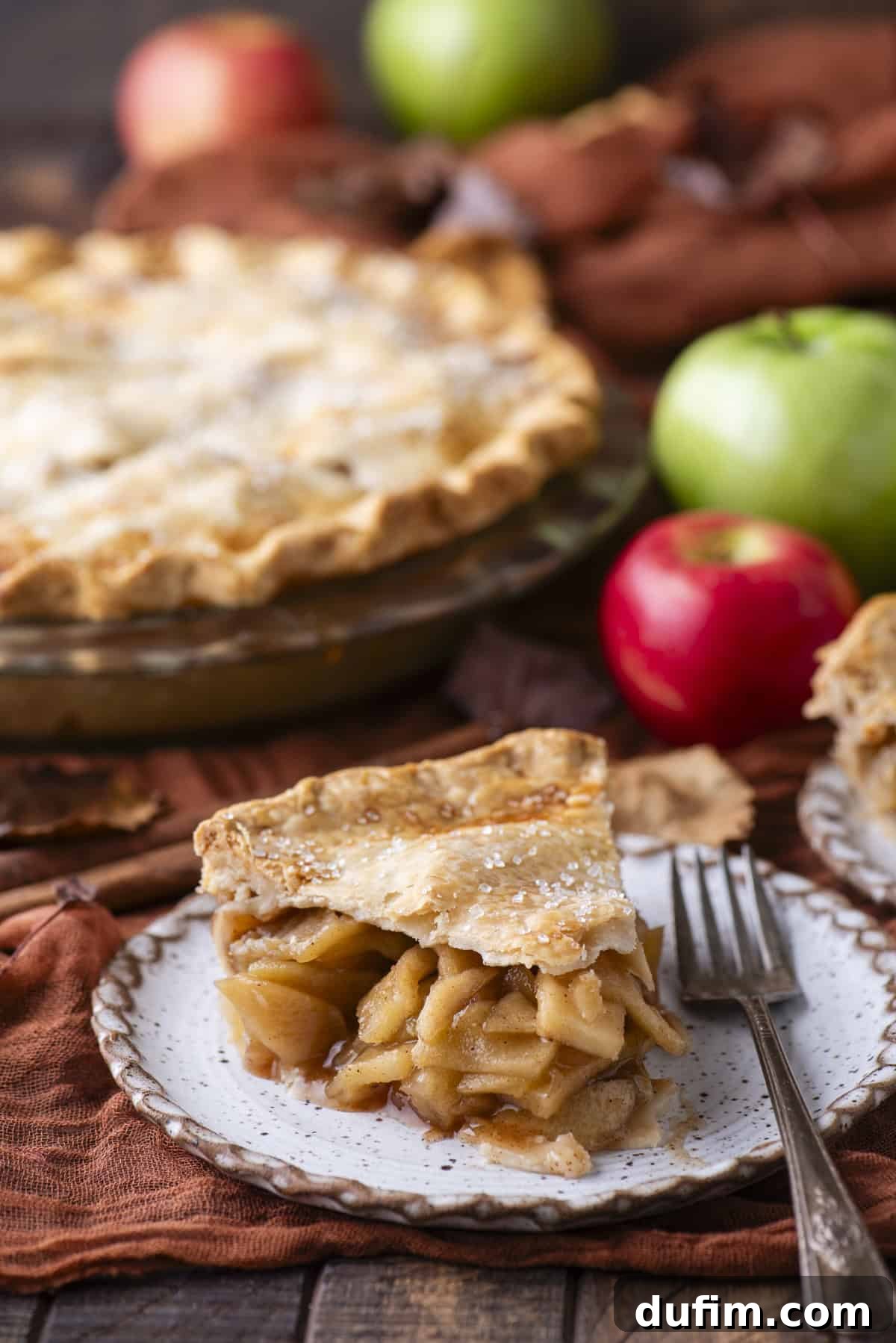 A perfectly baked slice of apple pie on a small white plate, accompanied by a fork. In the background, vibrant red and green apples are scattered, alongside a full, golden-brown apple pie, hinting at the abundance of fresh ingredients.