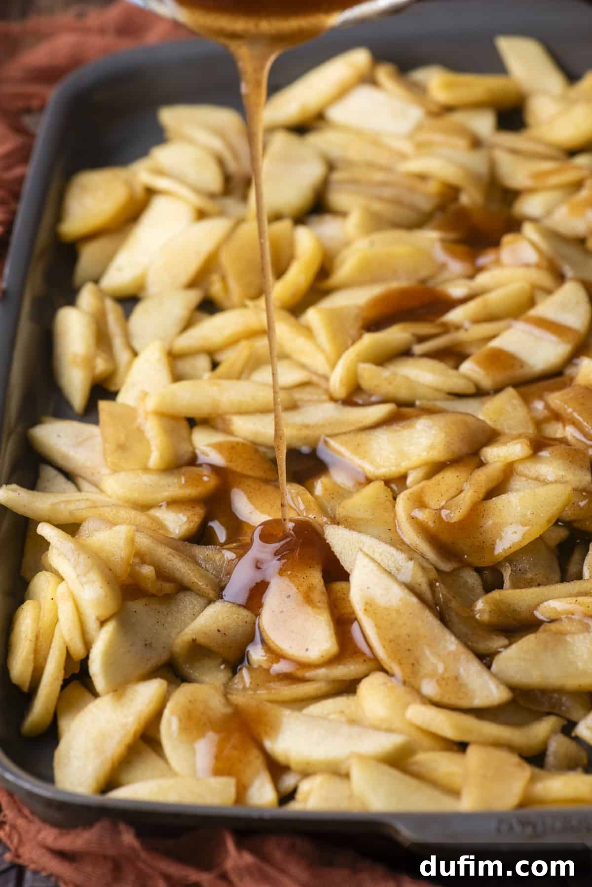 Cooked apple slices, evenly spread on a baking sheet, are being generously coated with a rich, glossy syrup poured from above, enhancing their flavor and texture.