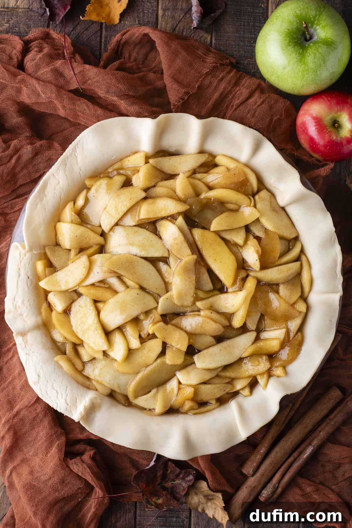 An inviting overhead view of an unbaked apple pie, nestled in its pie dish with a golden, uncooked crust, brimming with apple filling. It's elegantly surrounded by a colorful array of green and red apples, delicate fall leaves, and fragrant cinnamon sticks, ready for baking.