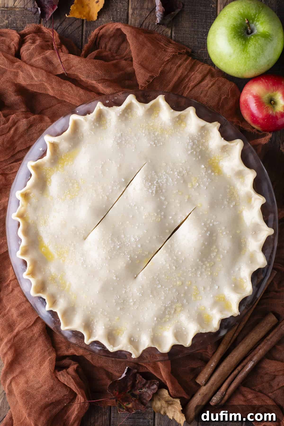 A stunning overhead shot of an unbaked apple pie, its top crust perfectly laid and crimped, awaiting its transformation in the oven. It is beautifully complemented by a scattering of fresh green and red apples, autumnal leaves, and cinnamon sticks, highlighting its homemade charm.