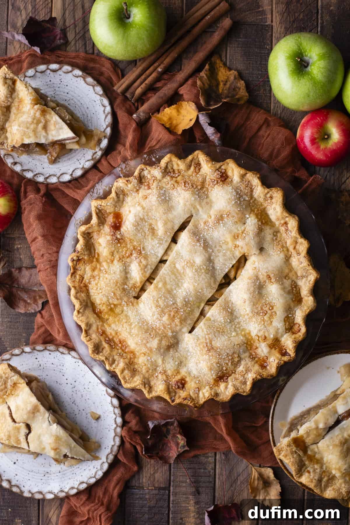 An enticing overhead view of a freshly baked apple pie, surrounded by individual slices of apple pie on small plates. Fresh green and red apples and scattered fall leaves complete the autumnal scene, highlighting the delicious outcome and readiness to serve.