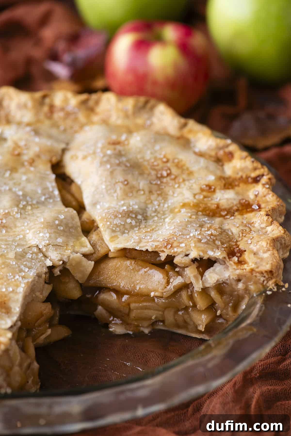 A delicious apple pie in a classic pie dish, with a single slice perfectly removed, revealing the rich, golden apple filling inside. In the background, a scattered array of fresh red and green apples suggests the quality of the ingredients used.