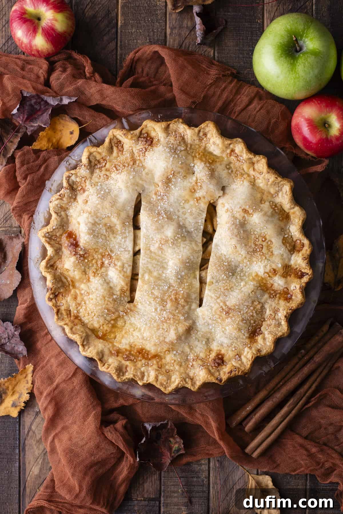 An inviting overhead view of a beautifully baked apple pie, artfully arranged on a rustic wooden surface. It is surrounded by an autumnal display of fresh green and red apples, scattered fall leaves, and fragrant cinnamon sticks, creating a cozy, harvest-inspired ambiance.