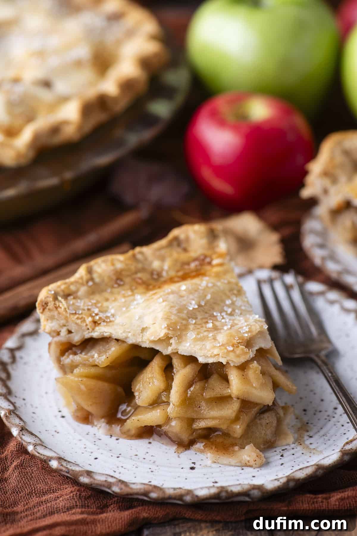 A delectable slice of apple pie served on a small white plate with a fork. The background showcases more fresh red and green apples, along with another portion of a full apple pie, highlighting the delicious outcome of the recipe.