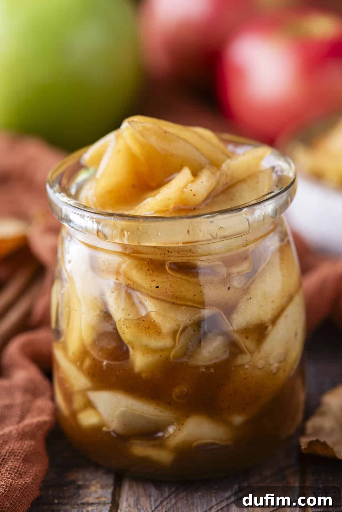 A small, elegant glass jar brimming with luscious apple pie filling, artfully displayed with vibrant red and green apples in the background.