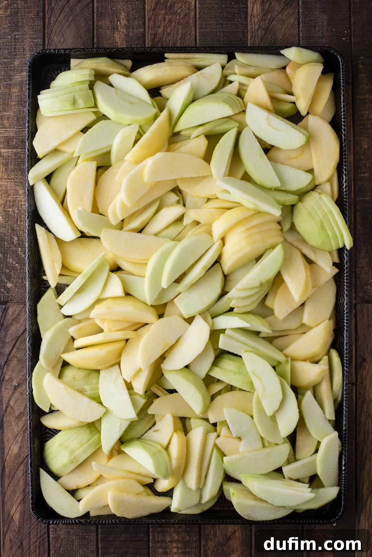 Overhead view of thinly sliced apples evenly spread across a baking sheet, ready for preparation.