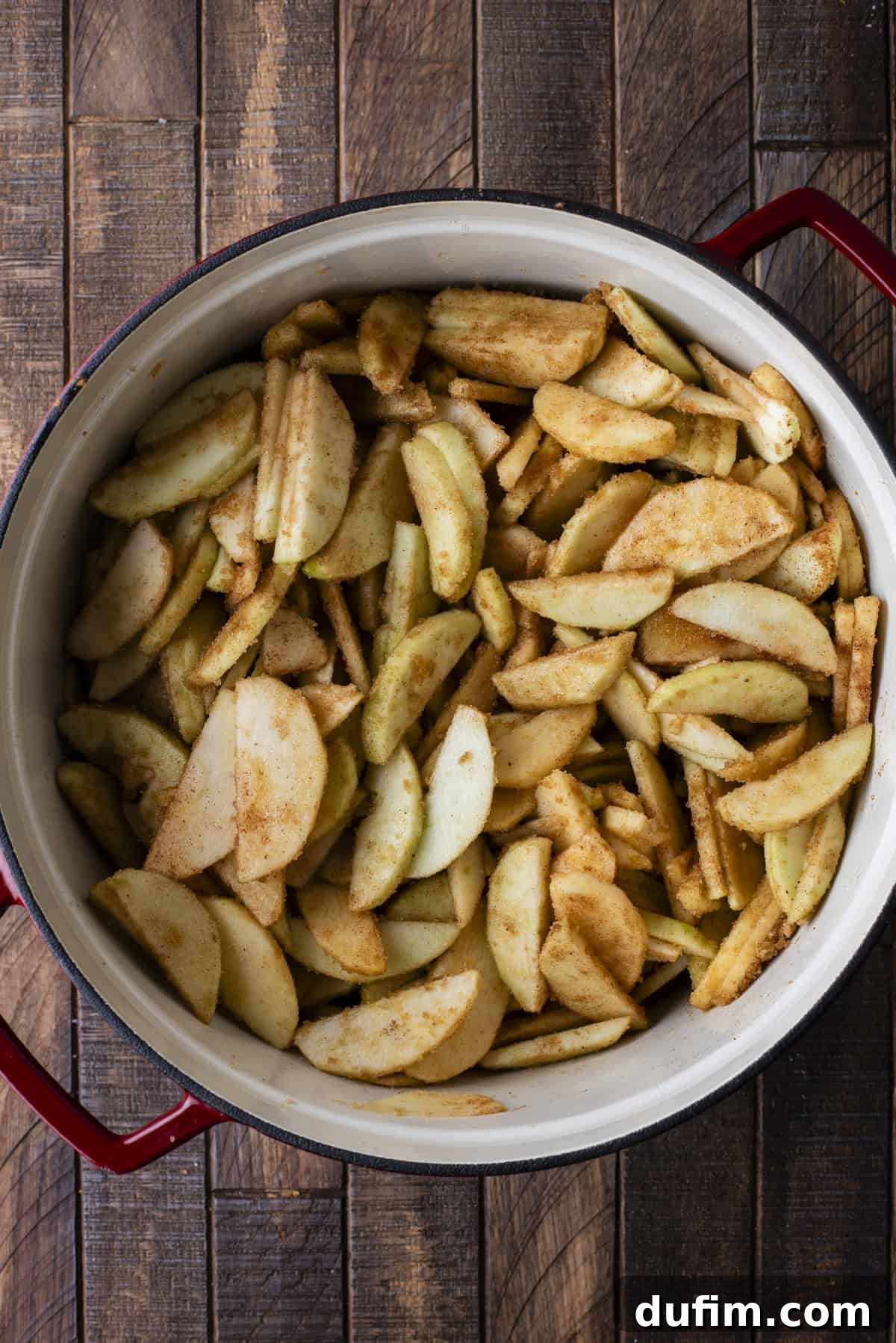 Overhead view of freshly sliced apples and a generous spice mixture in a large pot, resting on a rustic wooden surface, ready for cooking.