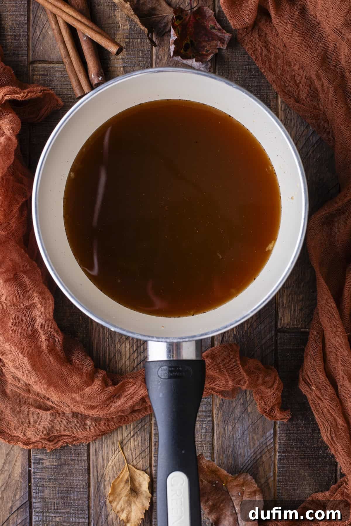Overhead view of a small saucepan filled with rich, simmering apple syrup, beautifully styled with autumn leaves and cinnamon sticks.