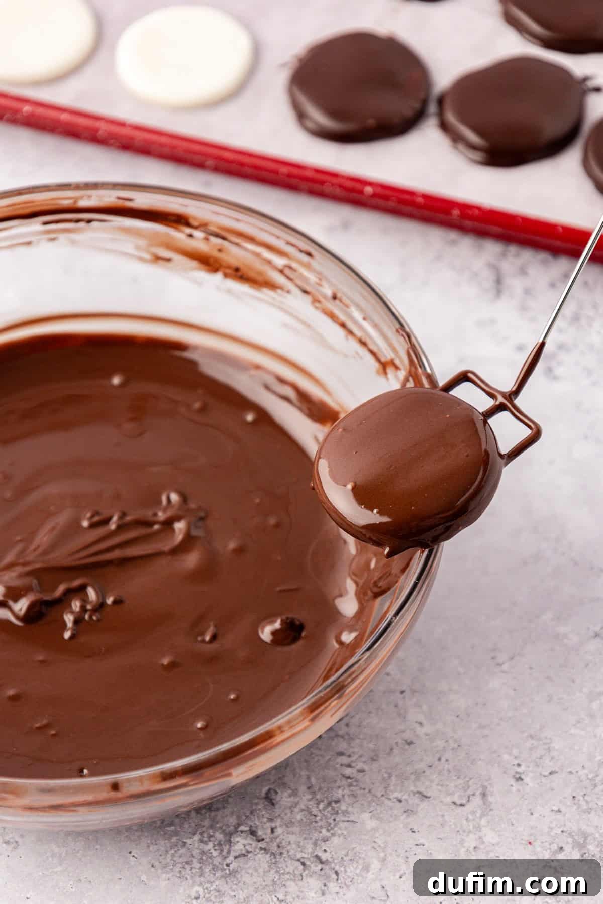 Cool & Creamy Homemade Peppermint Patties 13 A freshly dipped peppermint patty, still glistening with melted chocolate, being lifted gently from the edge of a clear glass bowl to allow excess chocolate to drip off.