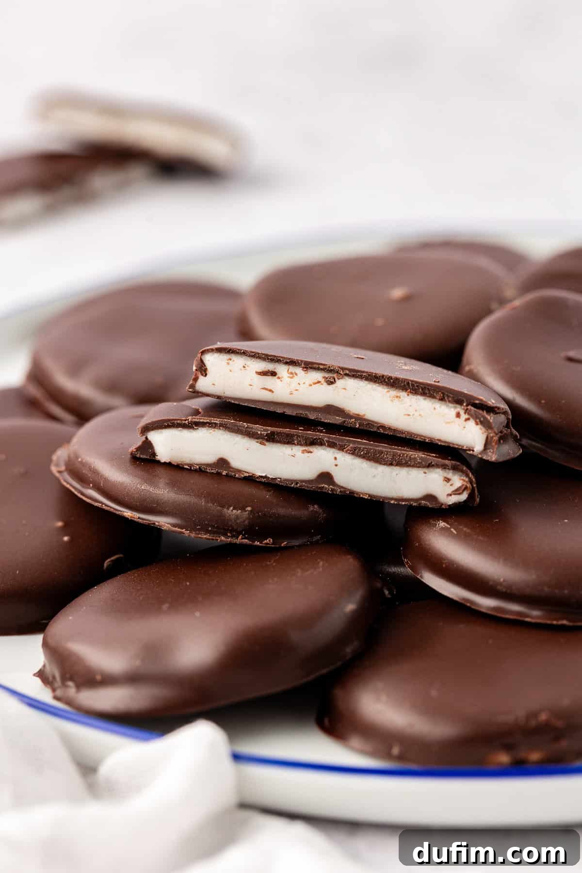 Cool & Creamy Homemade Peppermint Patties 3 A plate generously filled with whole peppermint patties, with one patty elegantly cut in half, showcasing its pristine white peppermint filling against the dark chocolate shell.