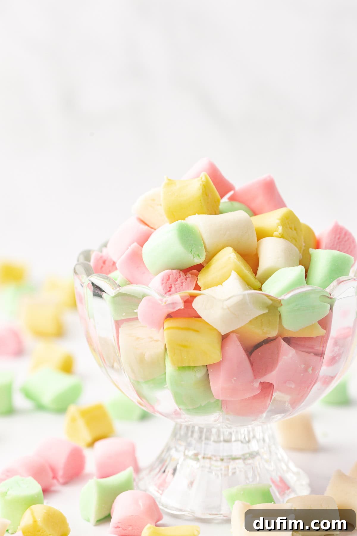 Melt-In-Your-Mouth Butter Mints 6 A glass trifle dish generously filled with a vibrant mix of white, green, pink, and yellow butter mints, with an inviting spill of more mints scattered around.