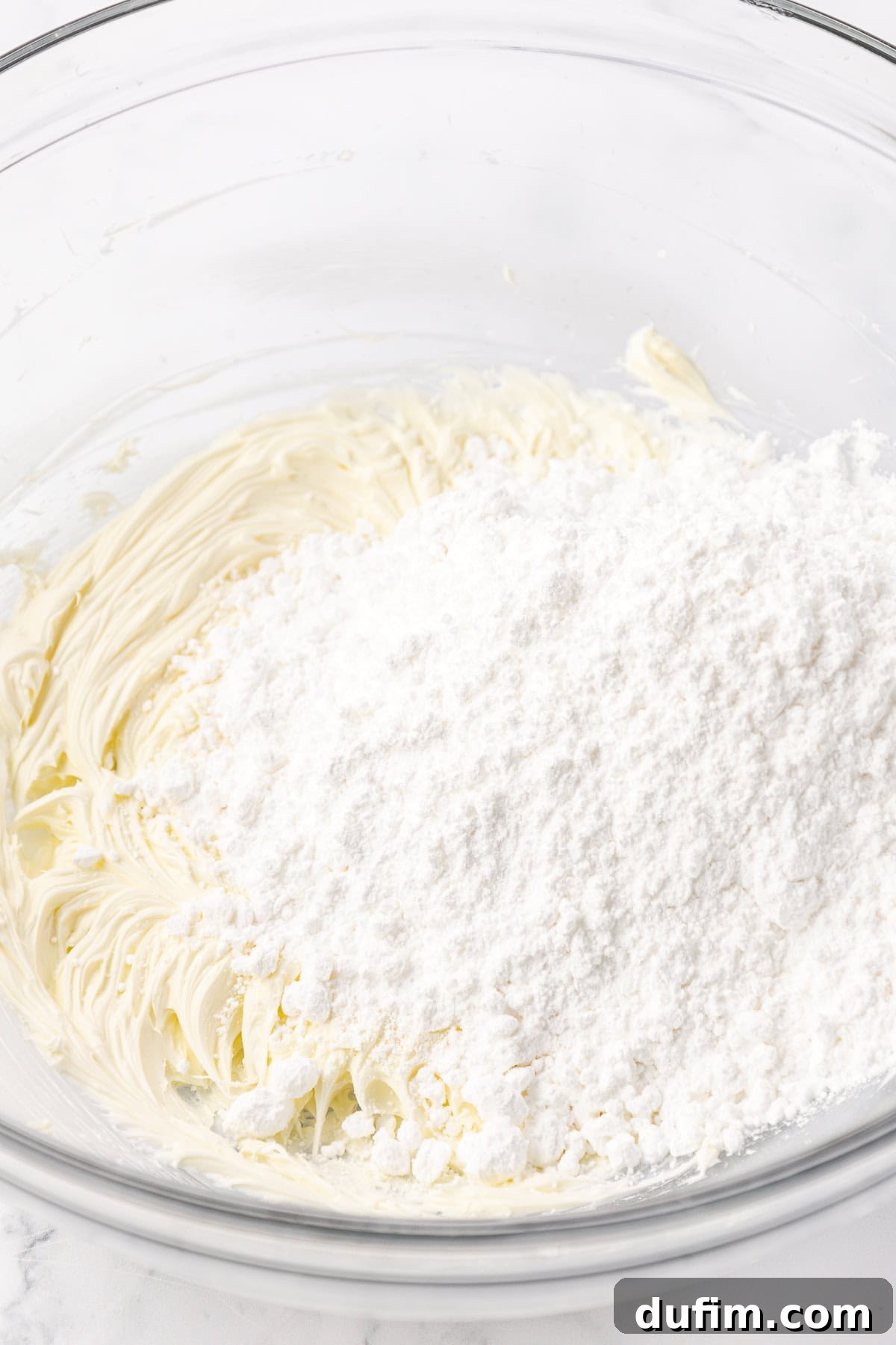 Melt-In-Your-Mouth Butter Mints 9 A clear glass bowl containing softened butter and a portion of powdered sugar, not yet combined, for the butter mint recipe.
