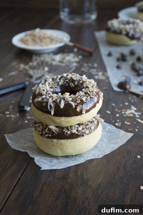 Close-up of baked coconut donuts with chocolate ganache and toasted coconut on a cooling rack.