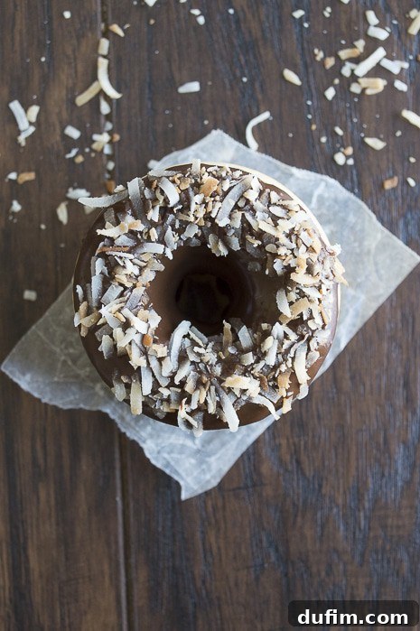 Freshly baked coconut donuts, some glazed with chocolate ganache and toasted coconut, arranged on parchment paper.