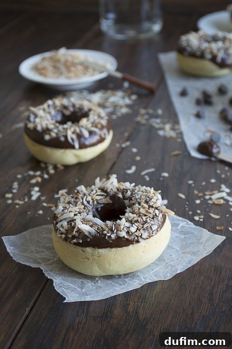 Multiple chocolate-glazed coconut donuts with toasted coconut scattered on a dark wooden surface, with a bowl of shredded coconut.