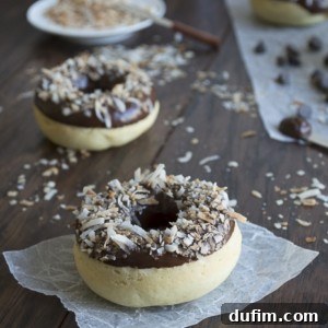 A chocolate-glazed coconut donut topped with toasted coconut sits on parchment paper, with more coconut donuts and a plate of shredded coconut in the background on a dark wooden surface.