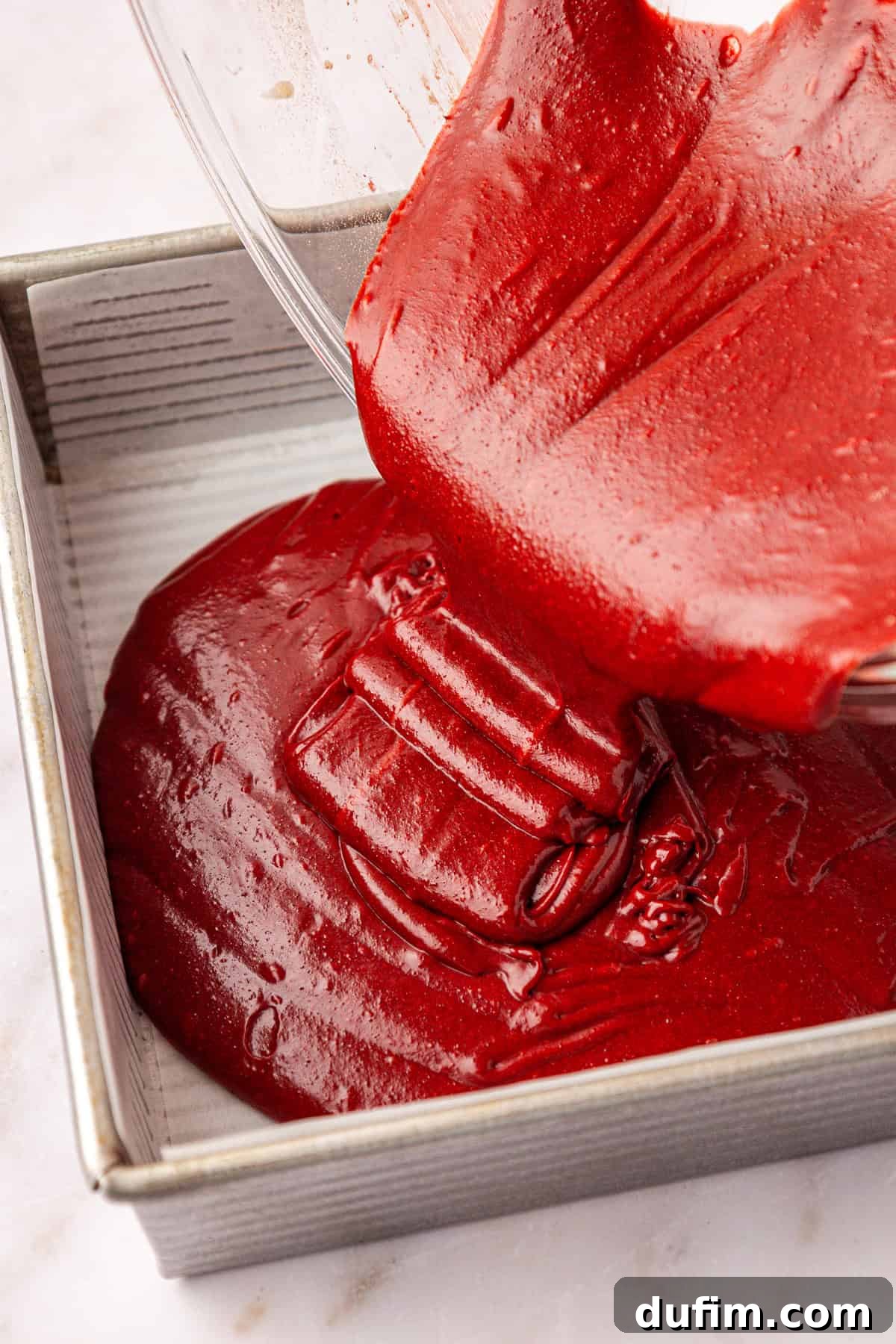 batter being poured out of a clear glass bowl into a square baking pan