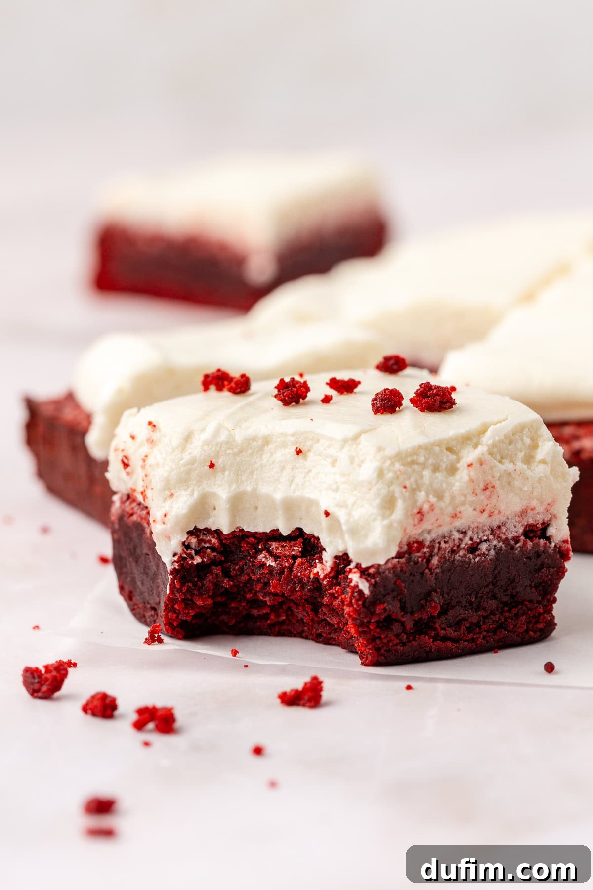 close up of a red velvet brownie topped with cream cheese frosting with a bite missing out of it, sprinkled with red velvet crumbs that are also scattered around