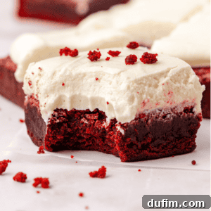 close up of a red velvet brownie topped with cream cheese frosting with a bite missing out of it, sprinkled with red velvet crumbs that are also scattered around