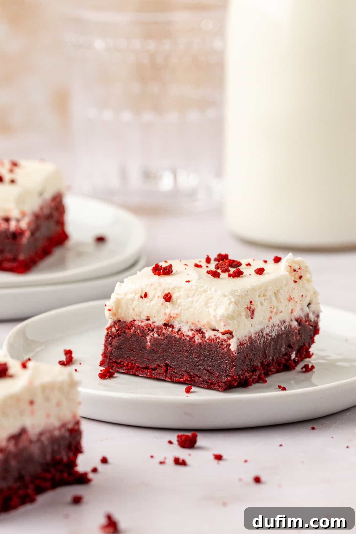 a red velvet brownie on a small white plate topped with cream cheese frosting and red velvet crumbs scattered around and on top of it