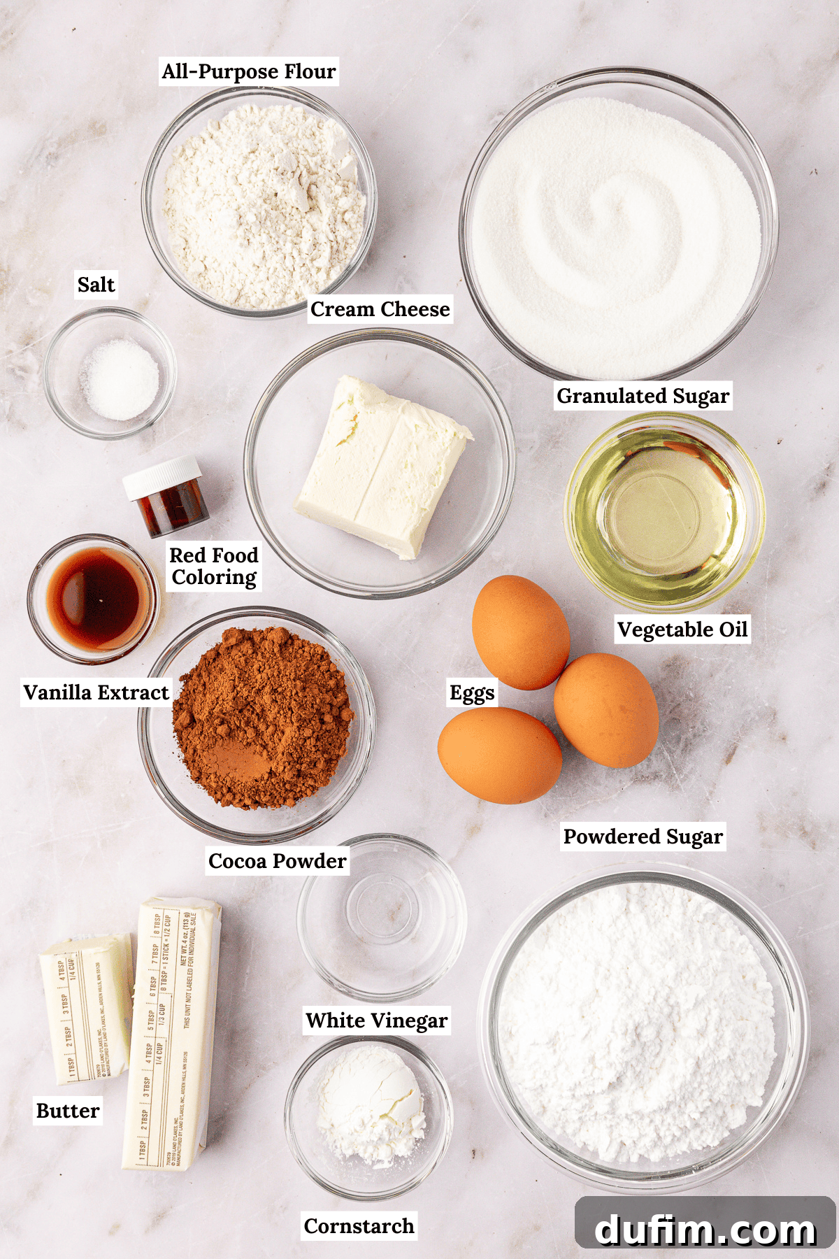 overhead view of the ingredients for red velvet brownies including all-purpose flour, granulated sugar, salt, cream cheese, red food coloring, vanilla extract, cocoa powder, eggs, vegetable oil, butter. white vinegar, cornstarch and powdered sugar
