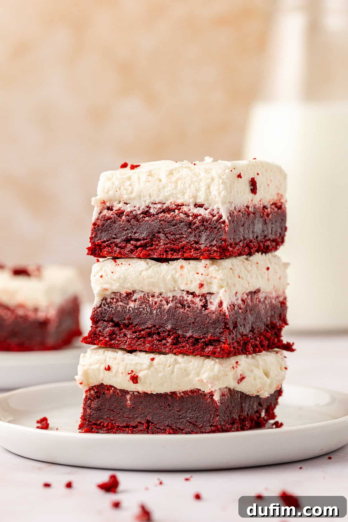 three red velvet brownies topped with cream cheese frosting stacked on a small white plate with red velvet crumbs scattered around