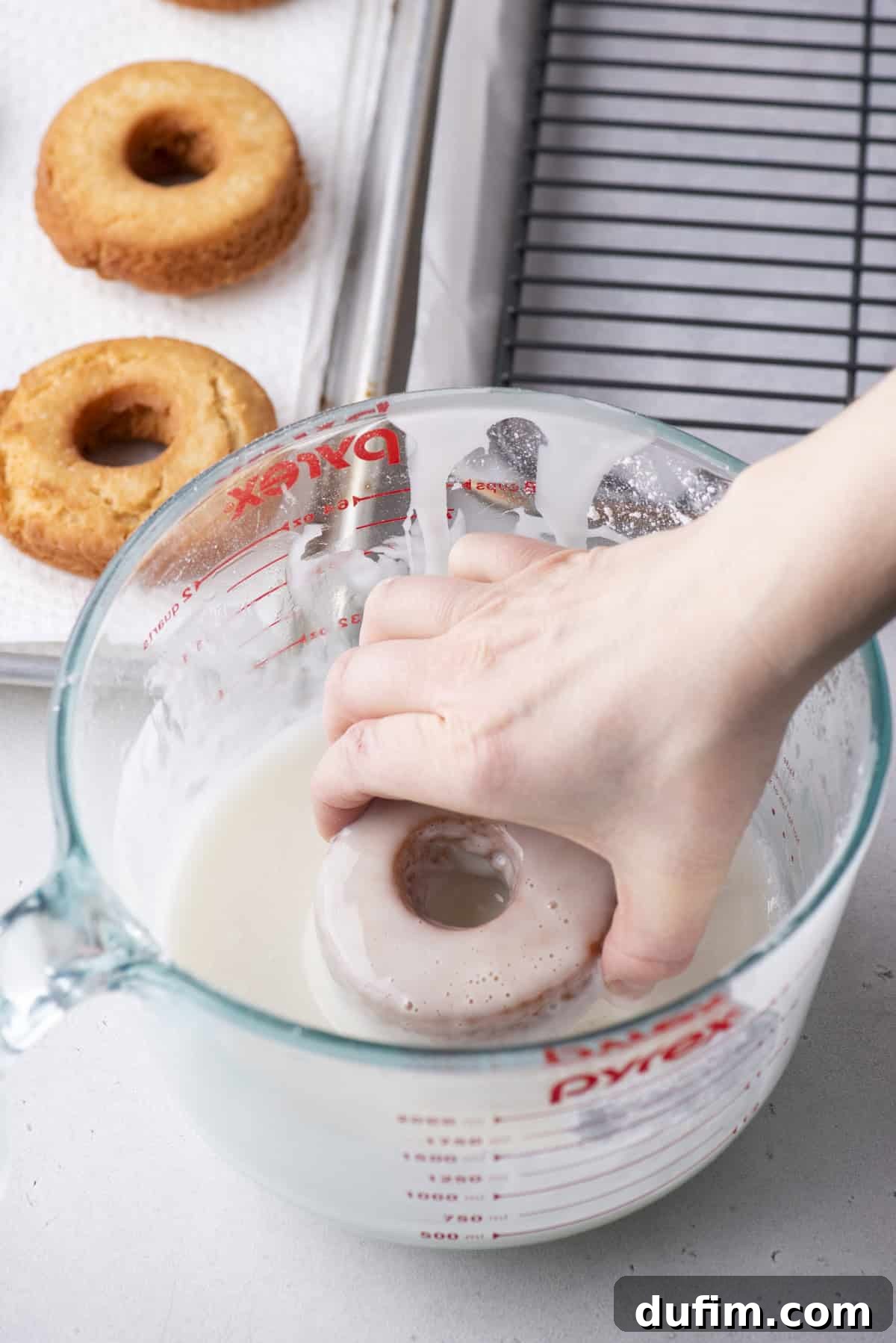Effortless Donut Glaze Perfection 13 A freshly dipped donut lifted from the glaze, with excess dripping back into the bowl, next to a cooling rack.