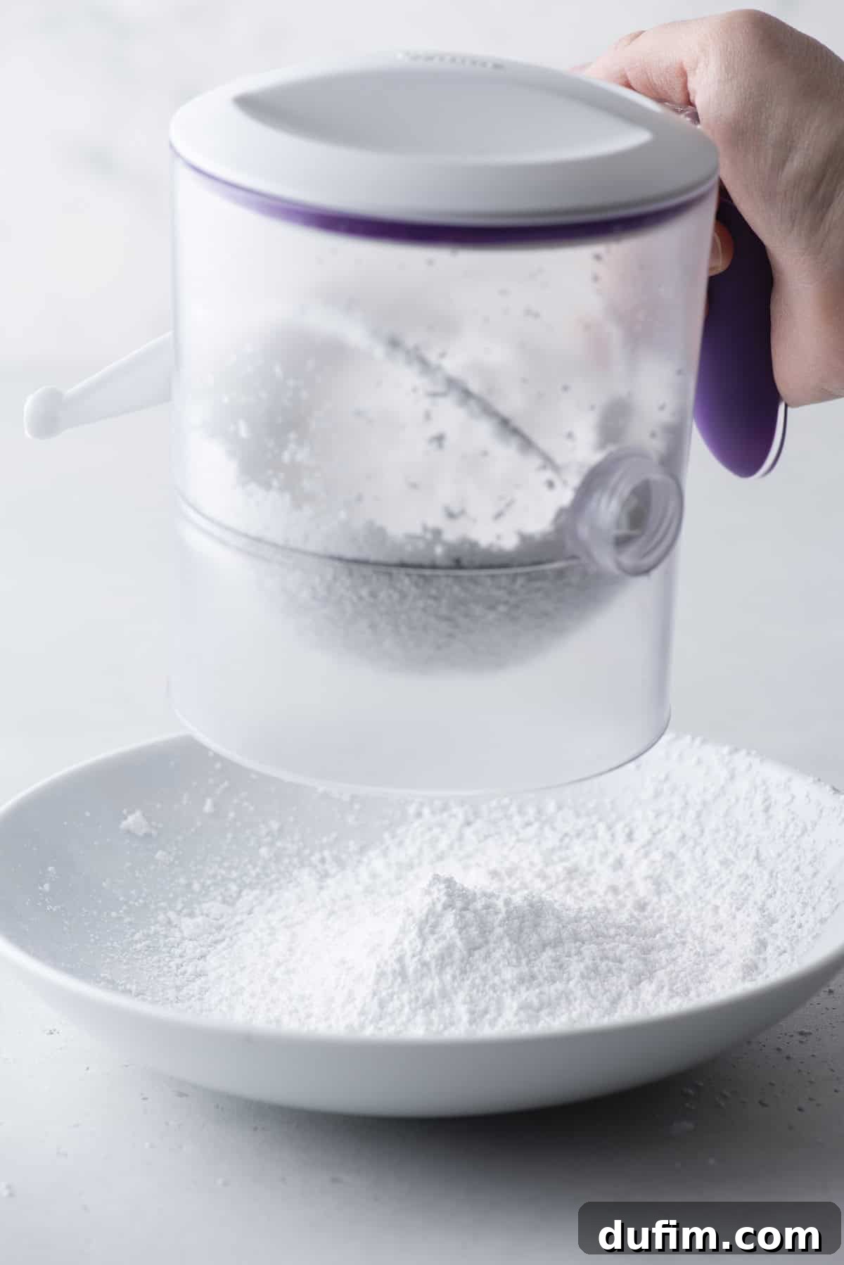 Effortless Donut Glaze Perfection 6 Powdered sugar being sifted through a fine-mesh sieve into a pristine white mixing bowl.