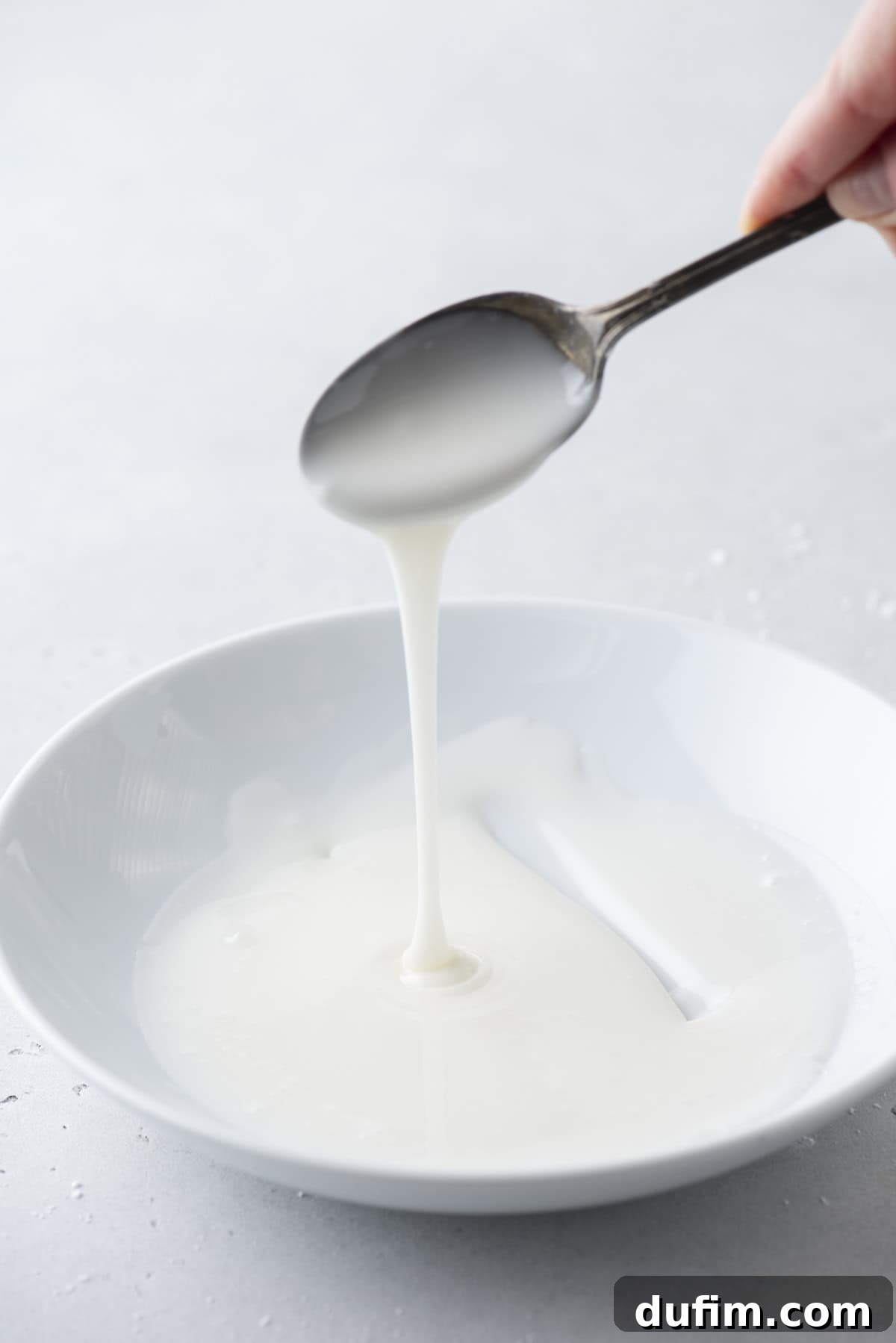 Effortless Donut Glaze Perfection 9 Finished donut glaze in a white bowl, with a spoon lifted above, demonstrating its smooth, pourable consistency.