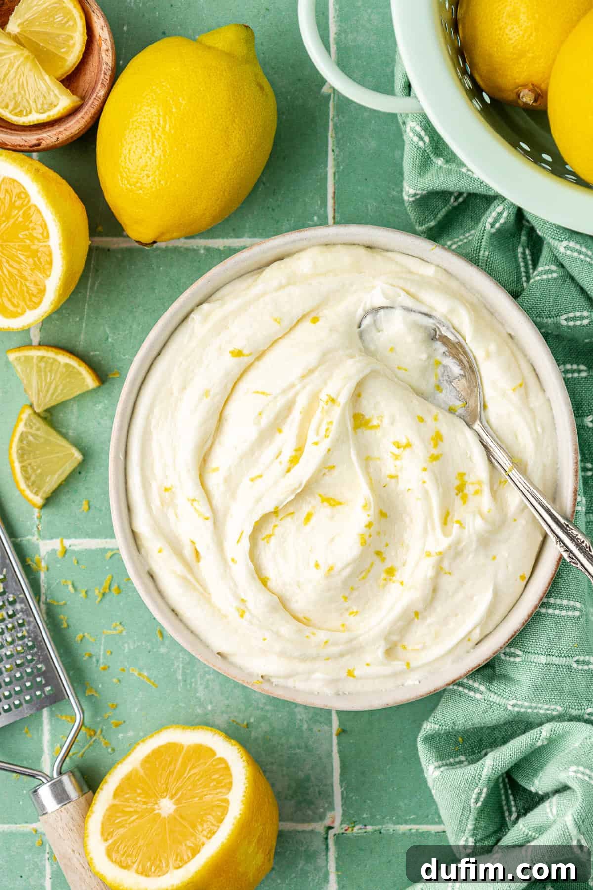 Luscious Lemon Cream Cheese Frosting 2 Overhead view of a bowl of vibrant lemon cream cheese frosting, garnished with fresh lemon zest and a spoon, surrounded by whole lemons, lemon slices, and a grater on a rustic surface.
