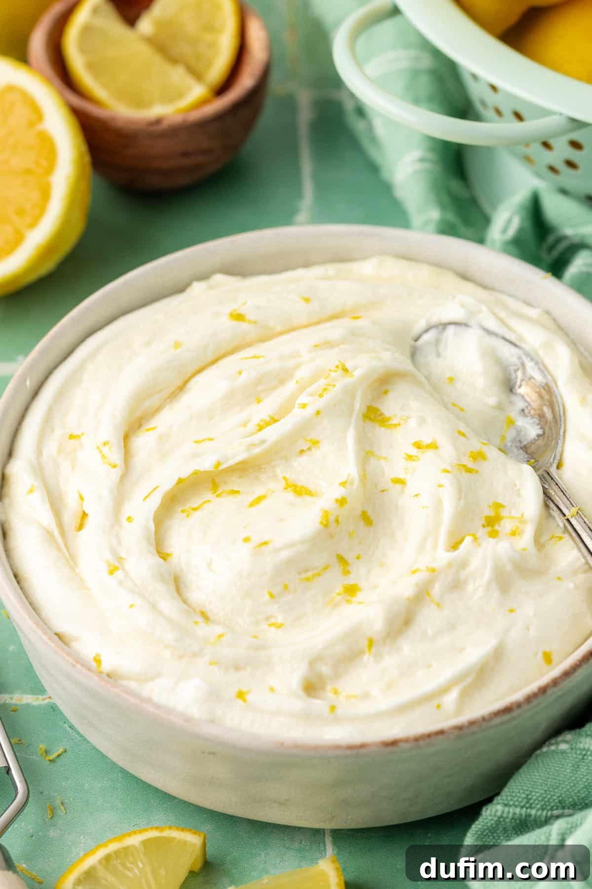 Luscious Lemon Cream Cheese Frosting 11 A pristine white bowl filled with creamy lemon cream cheese frosting, a spoon nestled within, garnished with fresh lemon zest and surrounded by bright lemon slices on a clean surface.