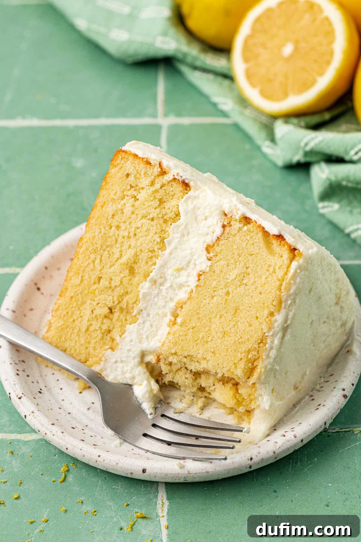 Luscious Lemon Cream Cheese Frosting 3 A perfectly frosted two-layer cake slice on a small white plate with a fork, elegantly placed against a blurred background of fresh lemons, highlighting the delicate texture of the lemon cream cheese frosting.
