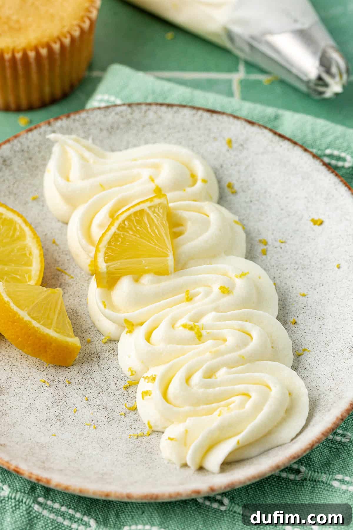 Luscious Lemon Cream Cheese Frosting 5 Beautifully piped lemon cream cheese frosting on a grey plate, adorned with vibrant lemon zest and a fresh lemon wedge. Two additional lemon wedges are artfully placed on the plate.
