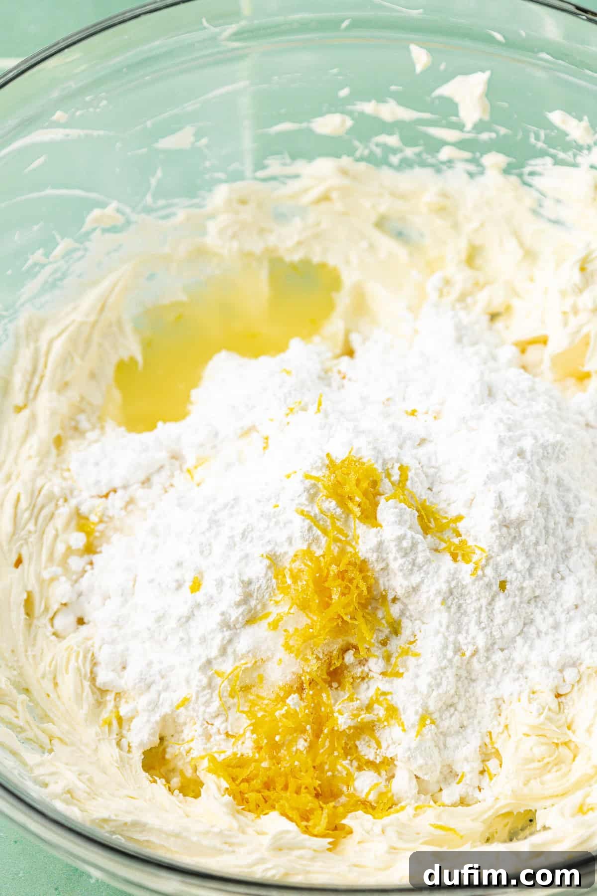 Luscious Lemon Cream Cheese Frosting 8 A clear glass bowl containing the key ingredients for lemon cream cheese frosting: creamed butter and cream cheese, powdered sugar, and lemon zest, ready to be combined.