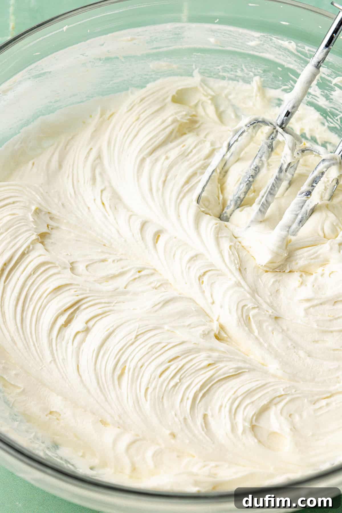 Luscious Lemon Cream Cheese Frosting 9 The fully mixed lemon cream cheese frosting in a large clear glass bowl, with an electric mixer paddle leaning in the creamy, smooth mixture, ready for use.