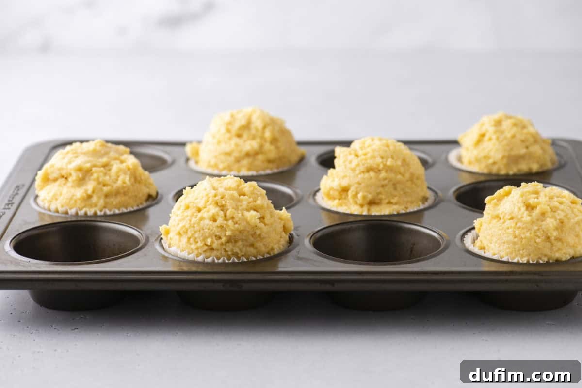 A muffin tray with seven scoops of uncooked corn muffin batter in paper liners, placed on a light gray countertop. The dough is rough and slightly mounded above the tray. The background is softly blurred.