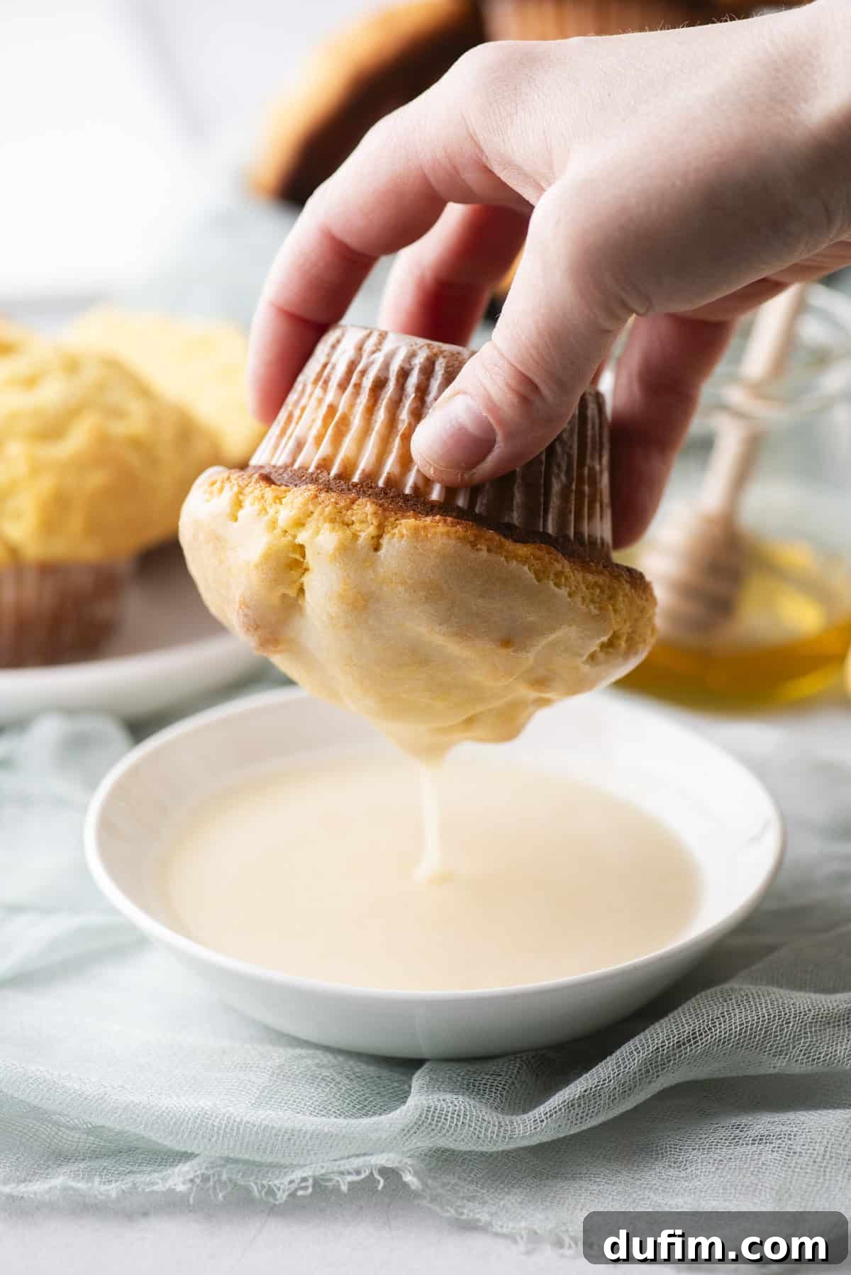 A hand dipping a muffin with a corrugated wrapper into a bowl of creamy glaze. More muffins are visible on a plate in the background, alongside a jar with a honey dipper. A soft, light blue cloth lies beneath.
