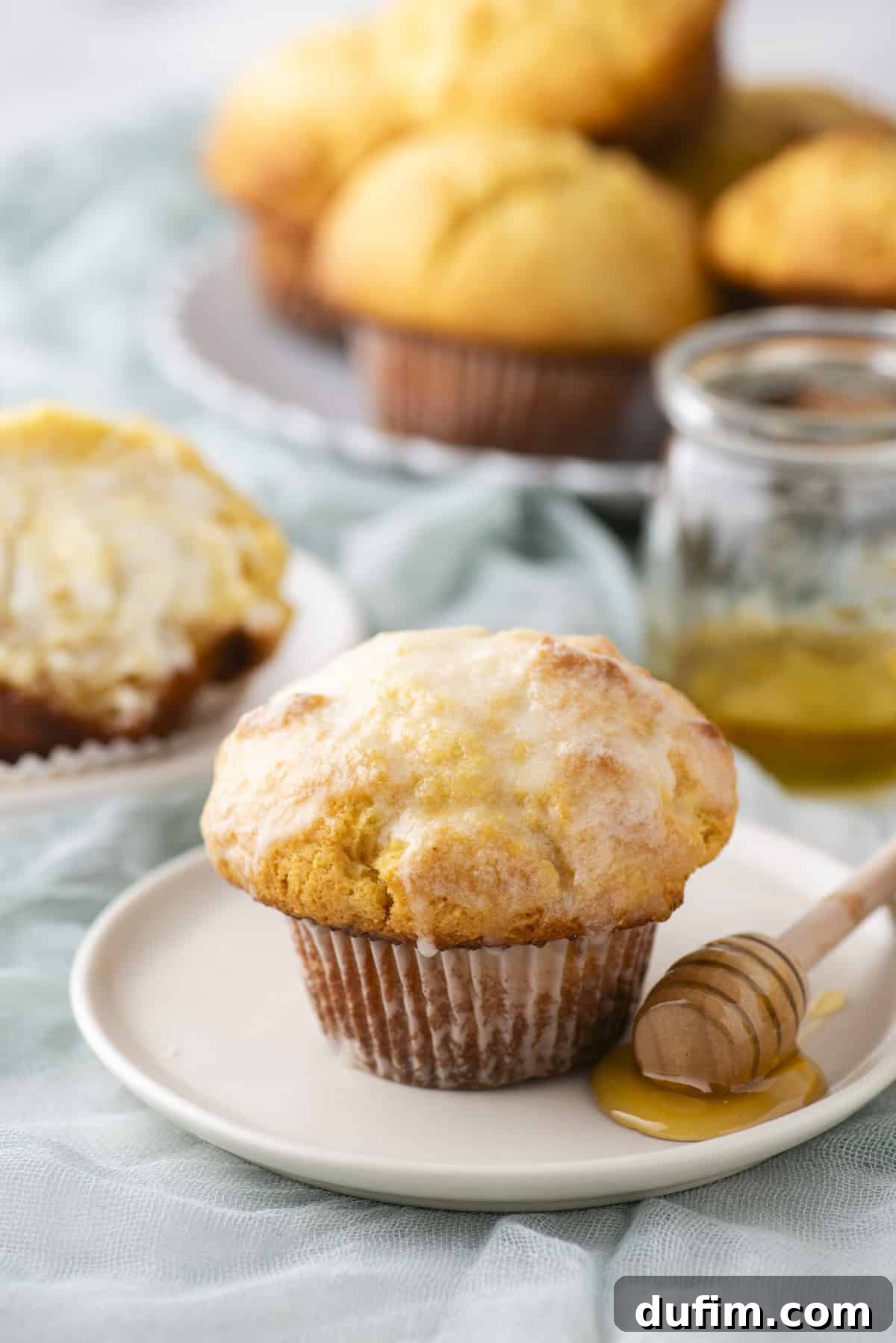 A muffin sits on a white plate with a honey dipper nearby. The muffin is golden brown with a light glaze. In the background, there's a partially eaten muffin on another plate and more muffins on a tray, with a jar of honey beside them.