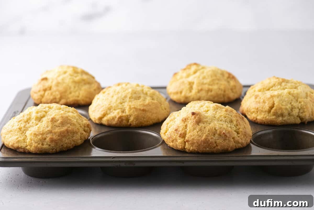 A muffin tray holds six golden-brown muffins, placed on a light-colored countertop. The muffins are slightly domed and appear freshly baked, with a soft, textured surface.