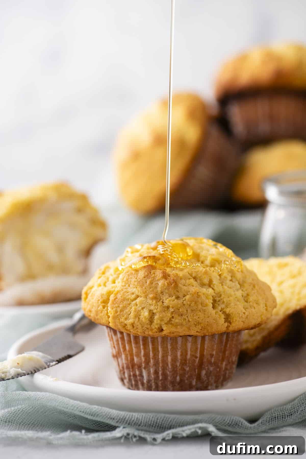 A muffin is on a white plate, being drizzled with honey. The background shows more muffins and a buttered muffin half. A small jar of honey is nearby. The scene is set on a light blue cloth and a marble surface.