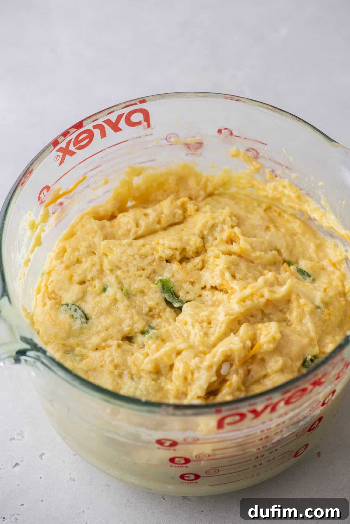 A clear measuring cup brimming with cornbread batter reveals bits of jalapeños mixed in, ready to be transformed into spicy corn muffins. The cup rests on a pristine white countertop.