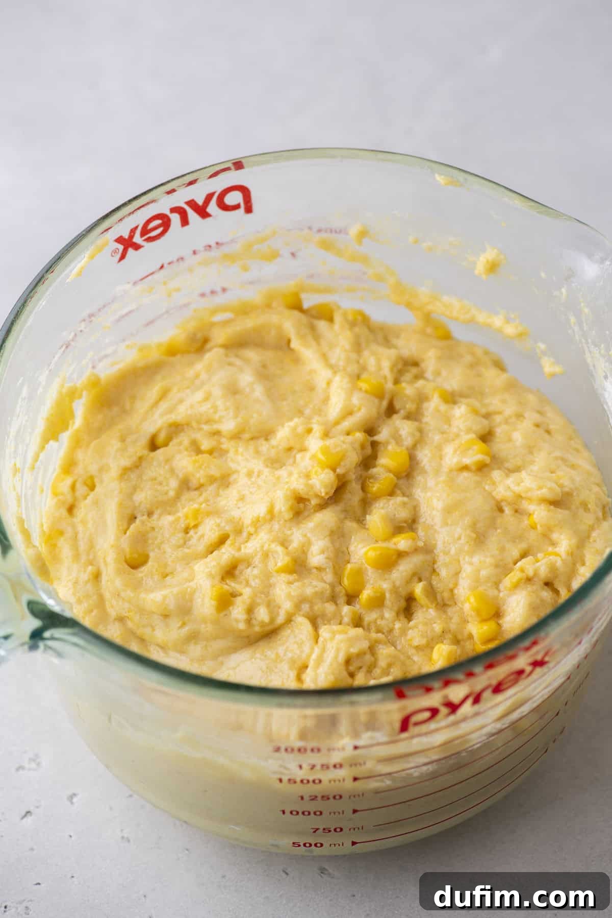 A glass Pyrex measuring cup brims with creamy yellow corn muffin batter, speckled with pieces of corn. Resting on a light-colored surface, the cup’s red measurement markings stand out against the glass.