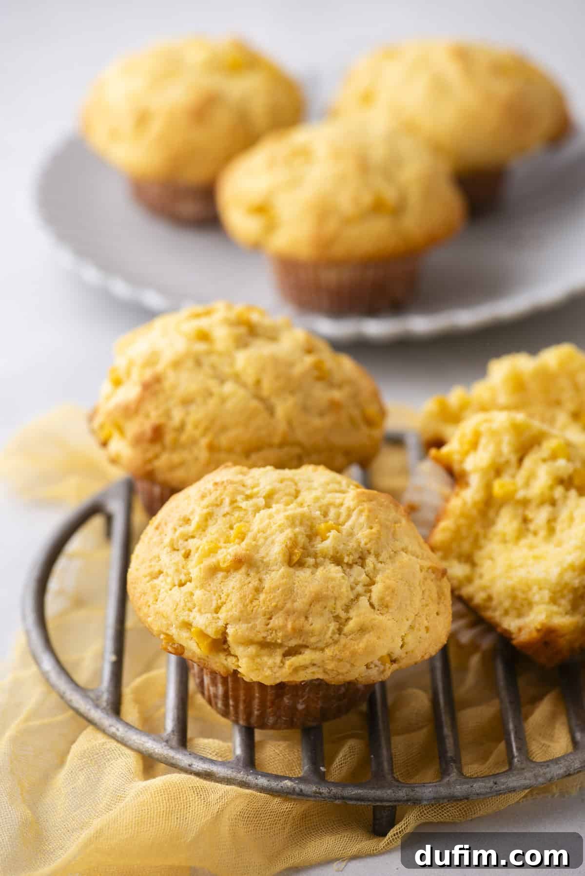 Freshly baked corn muffins rest on a cooling rack, with one split open to reveal its moist texture. A plate adorned with more corn muffins sits in the background, all set upon a light-colored cloth that adds to the inviting ambiance.