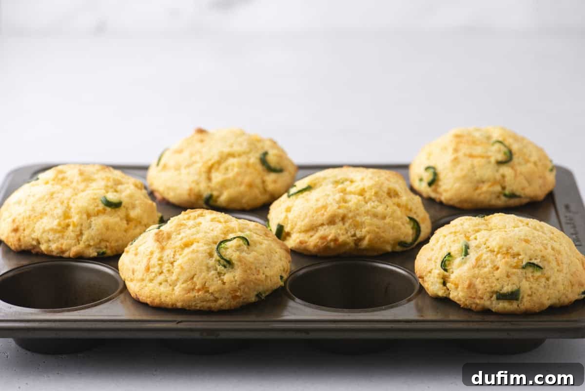 A muffin tin showcasing six freshly baked corn muffins, each adorned with slices of green jalapeños, sits invitingly. The golden brown corn muffins contrast beautifully against the light gray background.