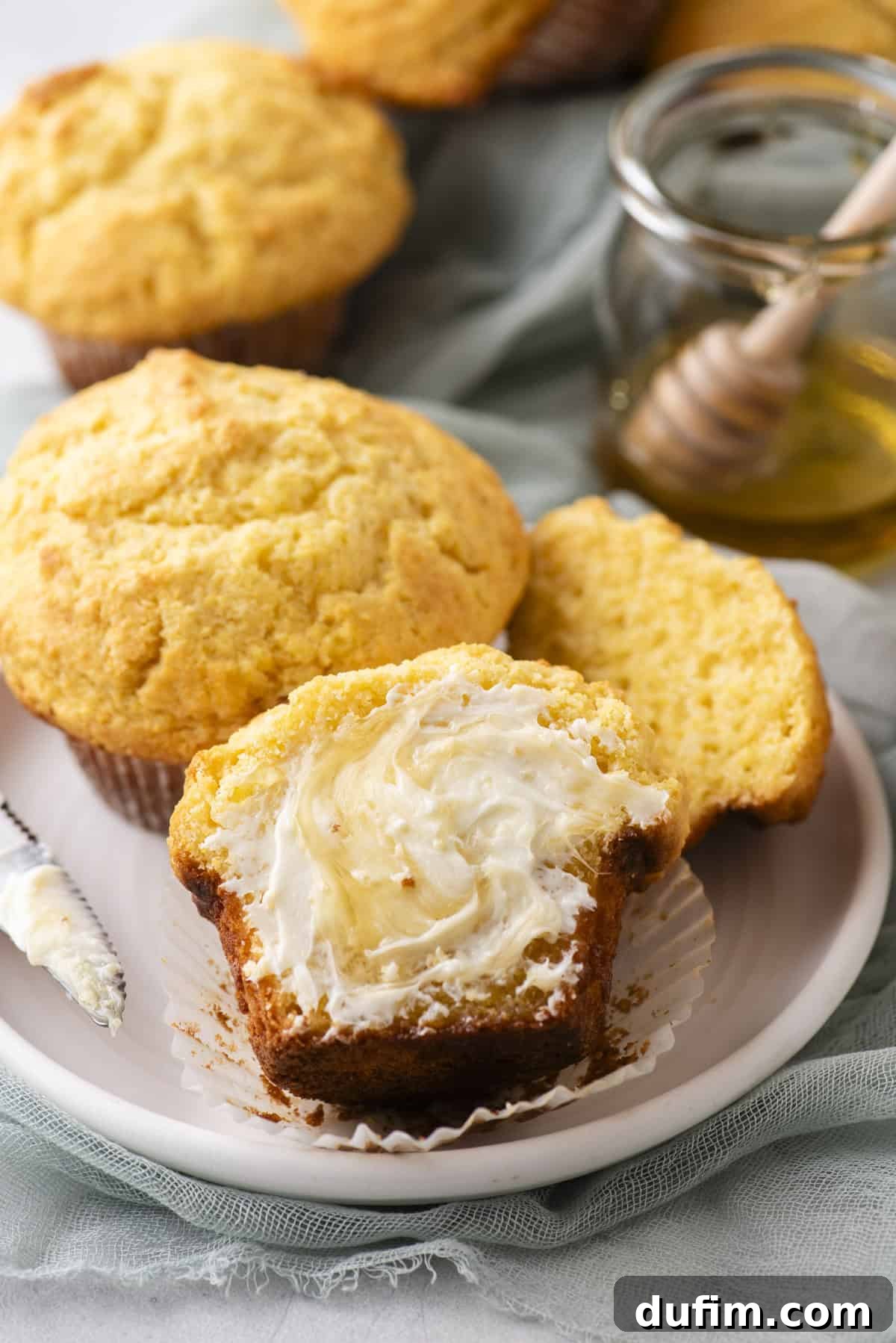 A plate with corn muffins, one cut open and spread with butter. A honey dipper rests on a jar of honey in the background. A green cloth is partially visible under the plate.