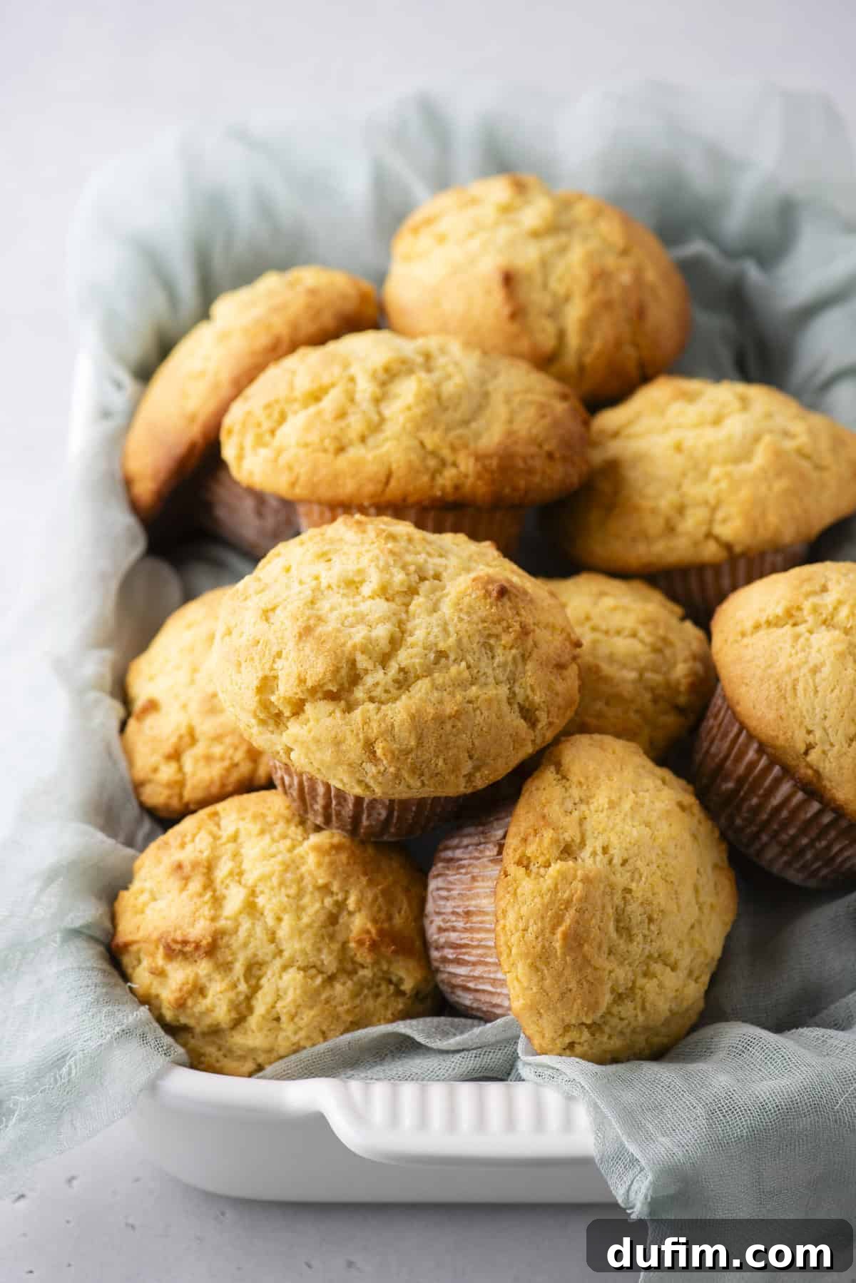 A white rectangular dish holds a stack of golden-brown muffins, lined with a light blue cloth. The muffins are fluffy with slightly cracked tops, suggesting they are freshly baked. The background is a smooth, light gray surface.