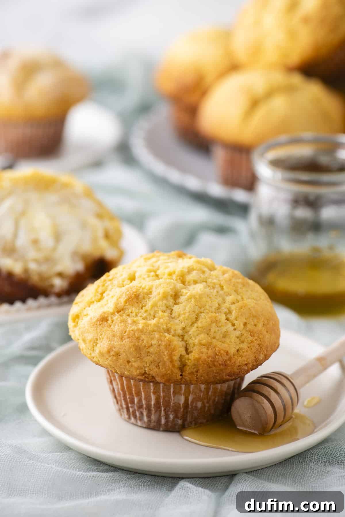 A cornbread muffin sits on a plate with a wooden honey dipper and honey drizzled nearby. In the background, more muffins are stacked on a plate, and one is cut open with butter. A jar of honey is also visible.